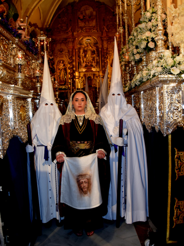 Una persona vestida con un hábito blanco y una capa con un rostro pintado, posando en una iglesia con un altar adornado con flores. La imagen transmite una sensación de solemnidad y tradición religiosa.