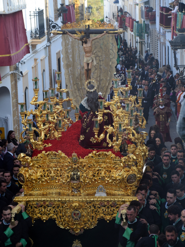 Procesión religiosa con paso ornado con oro y figura del Cristo en el centro, acompañada por un numeroso grupo de personas en trajes tradicionales.