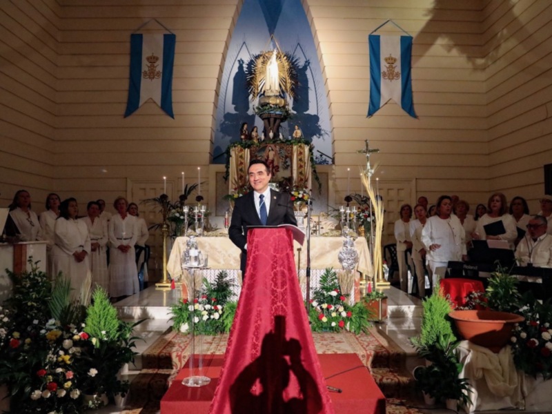 Celebración religiosa en una iglesia con un hombre en el altar y un coro en el fondo, adornada con flores y banderas.