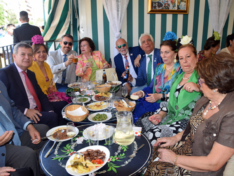 Una reunión formal en un ambiente festivo, con personas sentadas alrededor de una mesa decorada con platos de comida y bebidas, todos con vestimentas elegantes y accesorios de moda.