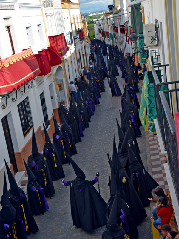 Procesión de la Semana Santa en una calle de Sevilla, con hermanos vestidos de luto y mantón rojo en las fachadas.