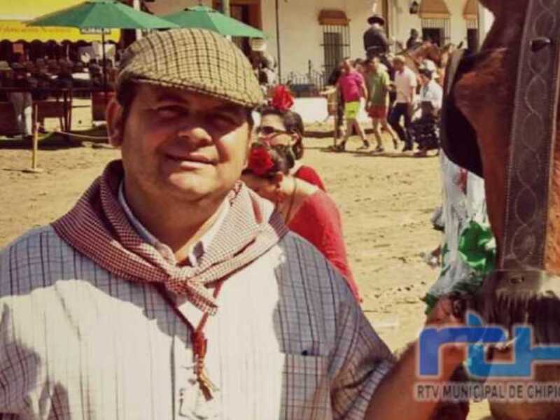 Un hombre en traje típico de la región de Chihuahua, México, posando para la cámara. El entorno muestra una celebración cultural con personas en el fondo y un caballo a la derecha.