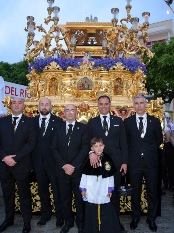 Celebración religiosa con hermandad y procesión, destacando un carro dorado y figuras religiosas.