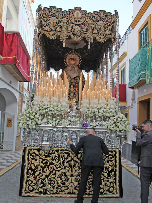 Procesión religiosa con estandarte ornado y velas iluminadas, en una calle de Sevilla.