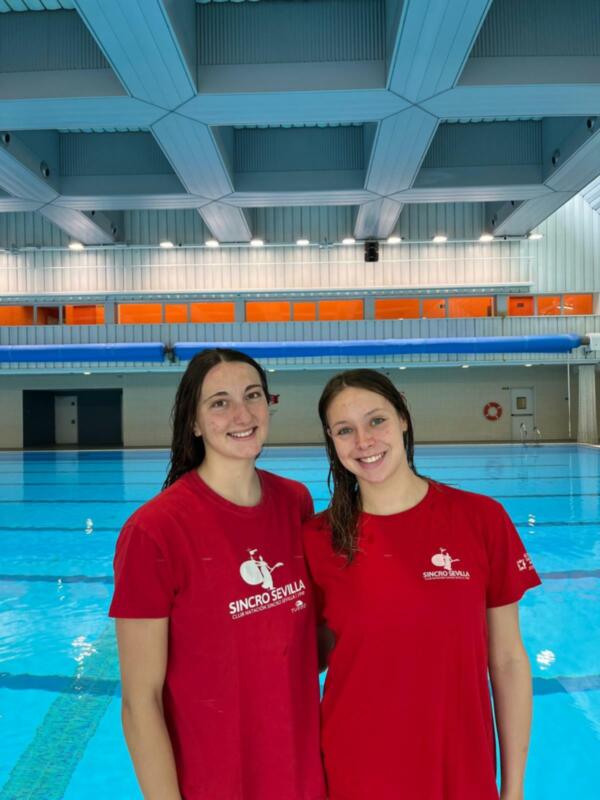 Dos nadadoras del Club Natación Sincro Sevilla sonríen frente a una piscina cubierta.