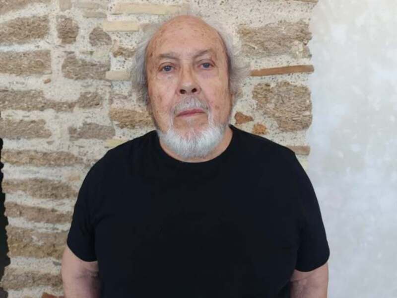 Un hombre con barba blanca y cabello gris, vestido con una camiseta negra, posando frente a una pared de ladrillo.