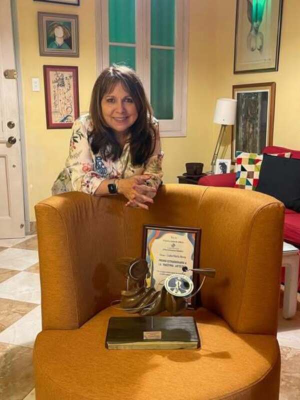 Una mujer sonriendo mientras sostiene un premio en una silla amarilla, con una decoración de arte en el fondo.