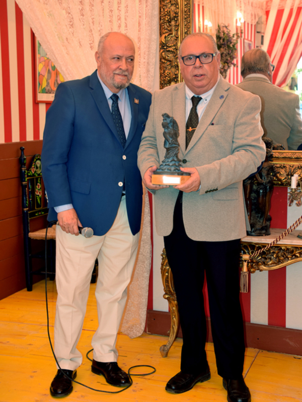 Dos hombres en trajes celebran un evento formal, uno sostiene un micrófono y el otro un trofeo en un salón decorado con cortinas y muebles antiguos.