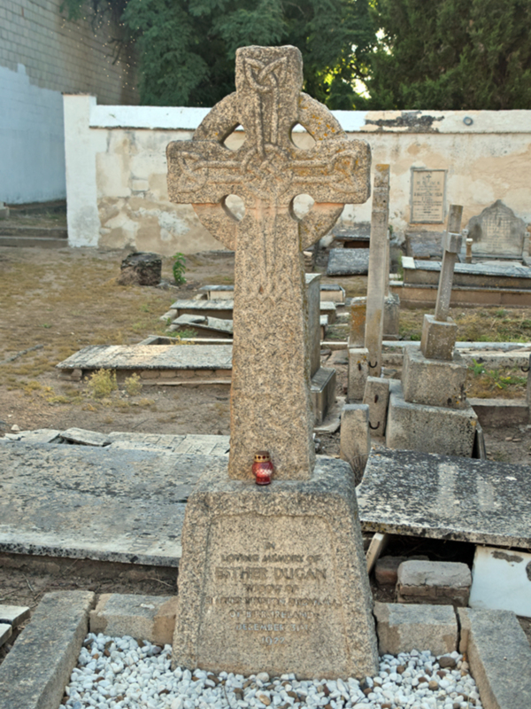 Un antiguo y delicado cruce celta en un cementerio, adornado con una lámpara de vela roja en la base, rodeado de otros monumentos sepulcrales.
