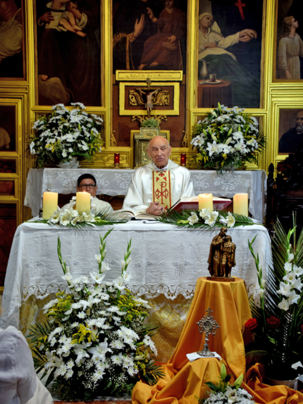 Un sacerdote celebra una misa en un altar adornado con flores y velas, bajo un retablo con pinturas religiosas.