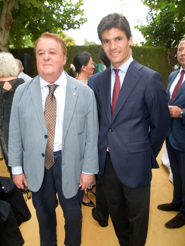 Dos hombres en trajes de negocios posan juntos en un evento al aire libre.