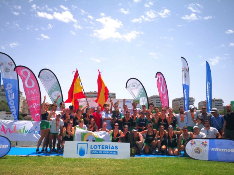 Equipo de remo celebrando con banderas y premios en un evento deportivo al aire libre.