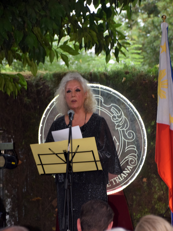 Mujer hablando en un evento al aire libre con un escudo y una bandera de fondo.