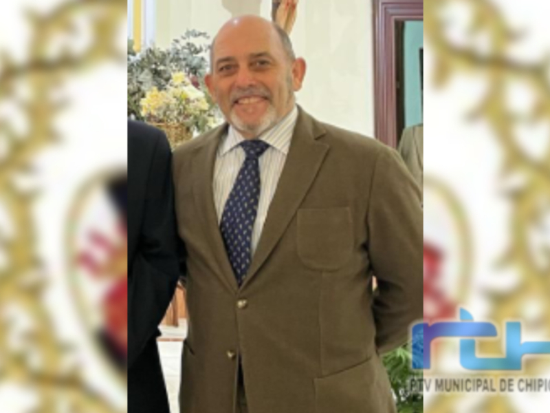 Retrato de hombre en traje, sonriendo, con el escudo de Chipiona en el fondo.