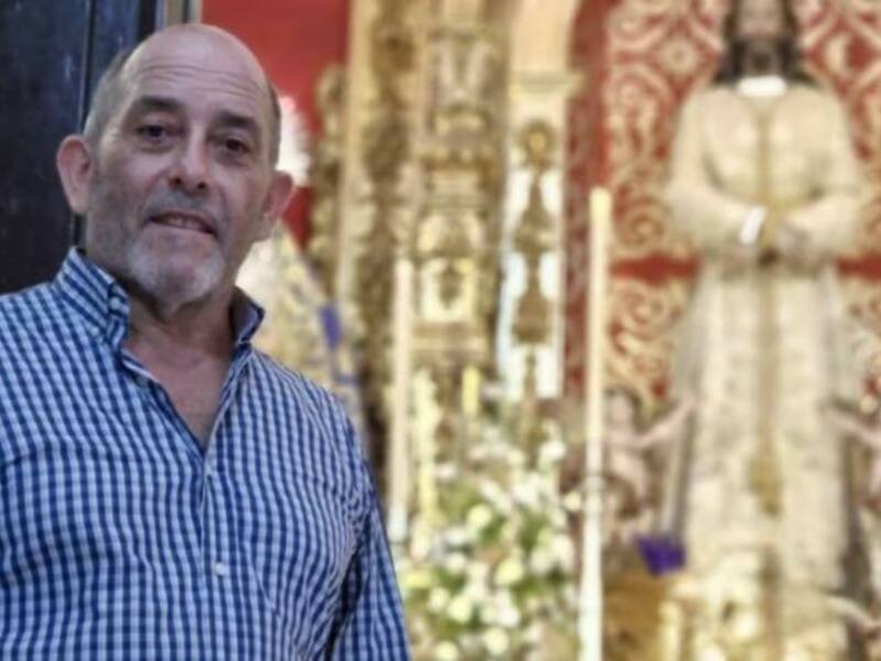 Un hombre con una camisa a cuadros azul y blanca, posando frente a una figura religiosa vestida de blanco, con detalles dorados y una corona en la cabeza, en un ambiente interior con decoraciones festivas. La imagen sugiere un evento cultural o religioso.