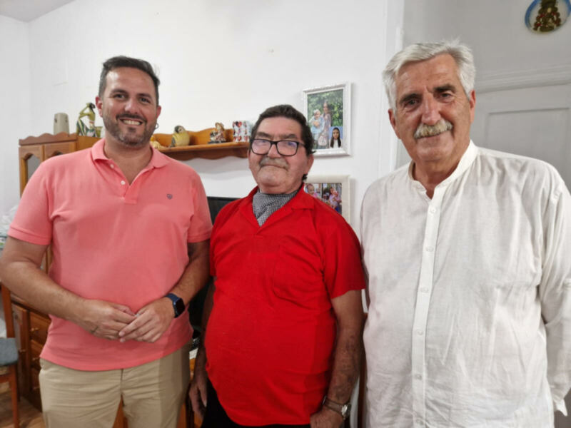 Tres hombres sonrientes en una sala de estar, rodeados de muebles y decoración familiar.