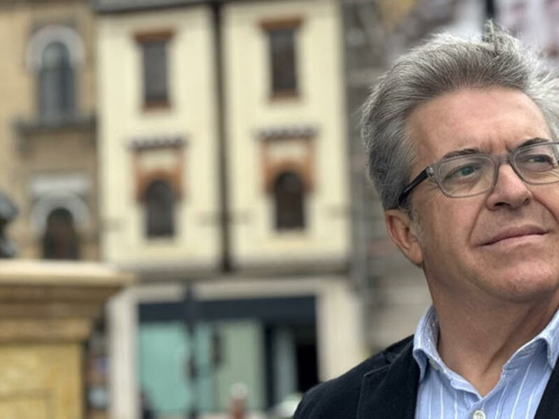 Hombre con gafas y chaqueta, frente a un edificio antiguo y una estatua en el fondo.