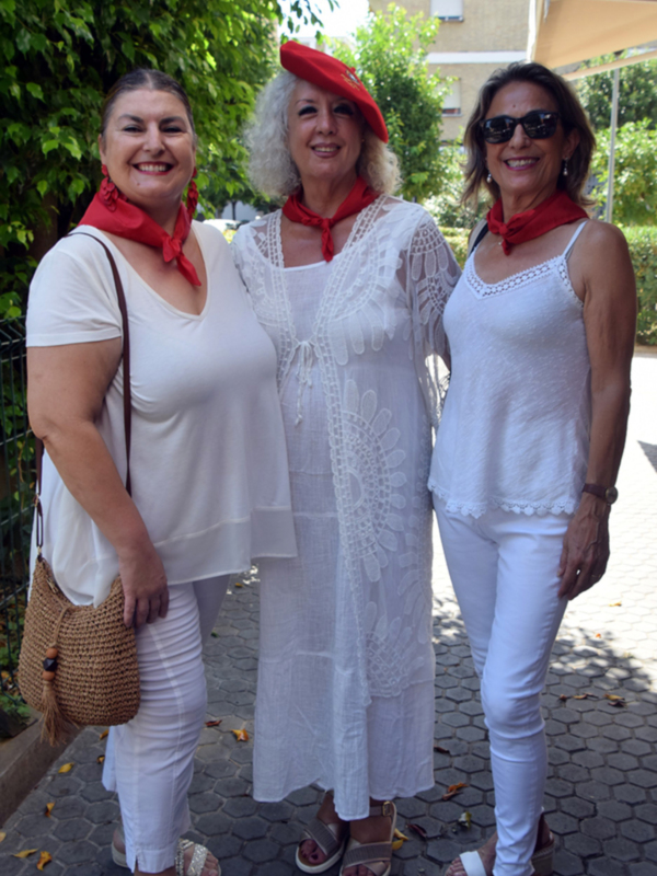 Tres mujeres sonrientes en un entorno urbano, vestidas con ropa blanca y accesorios rojos, posando para la foto. La escena está ambientada en una calle con vegetación y edificios en el fondo.