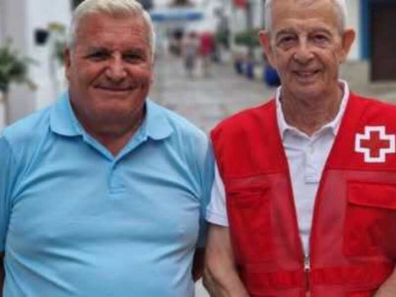 Dos hombres, uno con una chaqueta roja de la Cruz Roja, posan frente a un edificio blanco y un parque con flores.