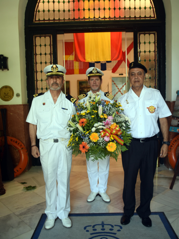Tres oficiales de la Armada española posan para una foto en el interior de un edificio con una bandera nacional en el fondo.