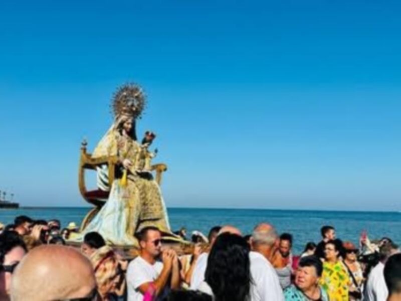 Procesión marítima con estatua procesional en la playa, bajo un cielo azul claro.