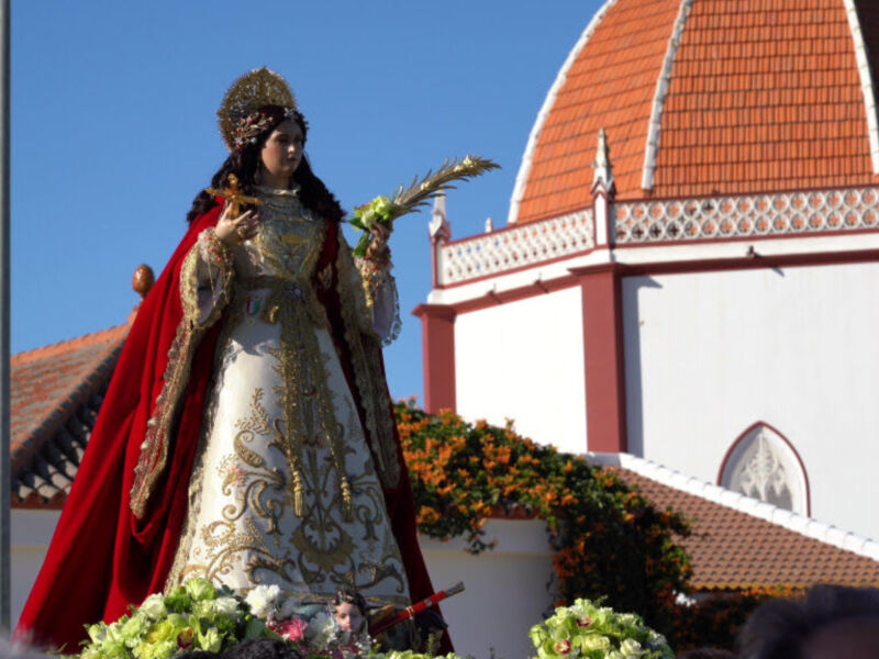 Estatua religiosa en procesión, vestida con traje tradicional y coronada, frente a una iglesia con cúpula roja y blanca, bajo un cielo azul claro.
