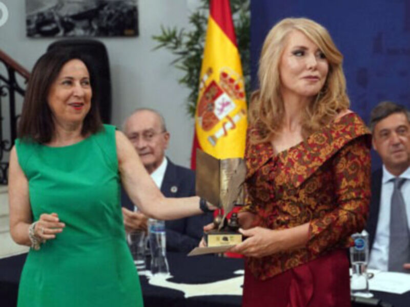 Una mujer con un vestido rojo sostiene un trofeo mientras otra mujer en verde observa en un evento formal con bandera española en el fondo.