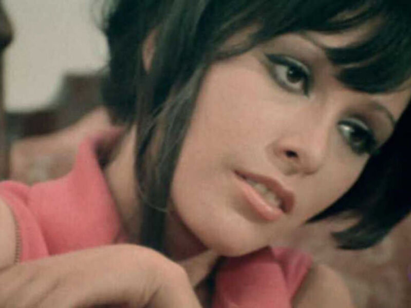 Una mujer con maquillaje de ojos oscuro y un vestido rojo, posando con una expresión seria.