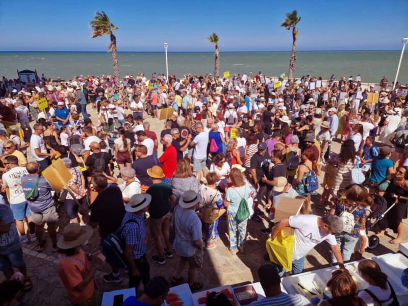 Una multitud de personas reunidas en la playa, algunos con pancartas y otros con sombreros y ropa de verano, disfrutando del clima soleado y el mar en el fondo.