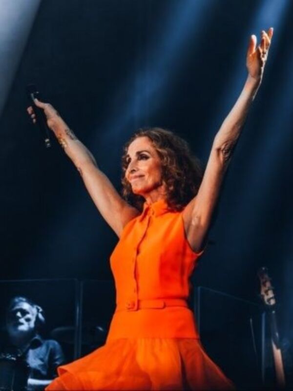 Una mujer con un vestido naranja brillante levanta las manos en un escenario con luces de fondo.