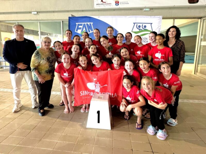 Equipo de natación sincronizada posando con trofeo y bandera en el fondo.