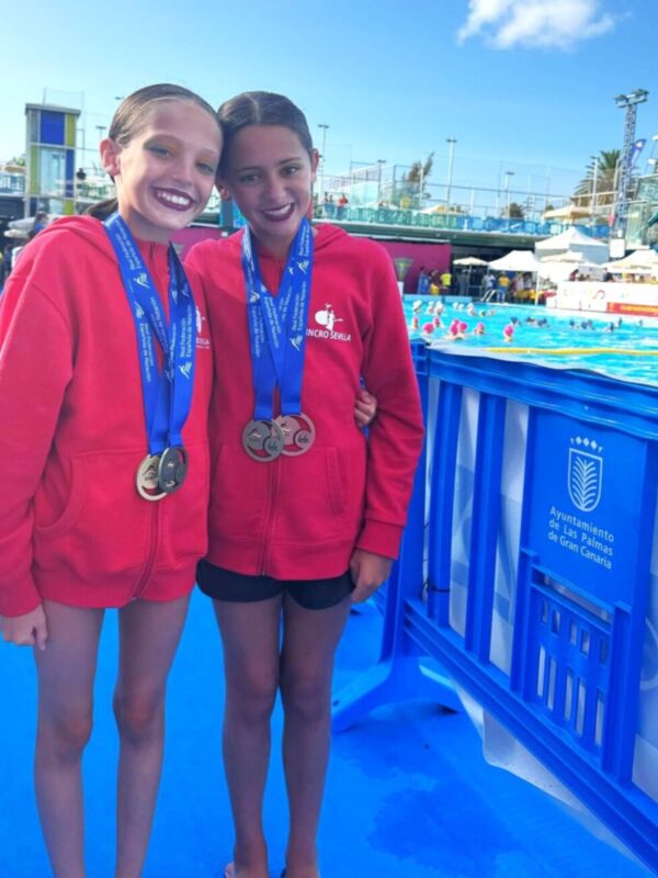 Dos nadadoras posan con sus medallas en una piscina al aire libre, con palmeras y edificios al fondo.
