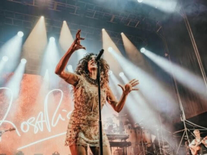 Mujer cantando en un concierto con luces de fondo y un panel con su nombre en la pared.