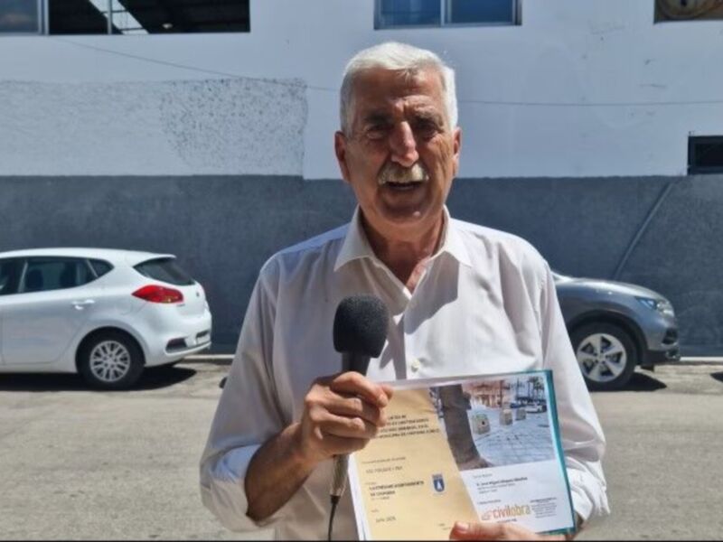 Hombre sosteniendo un certificado frente a un fondo de vehículos y un edificio blanco.