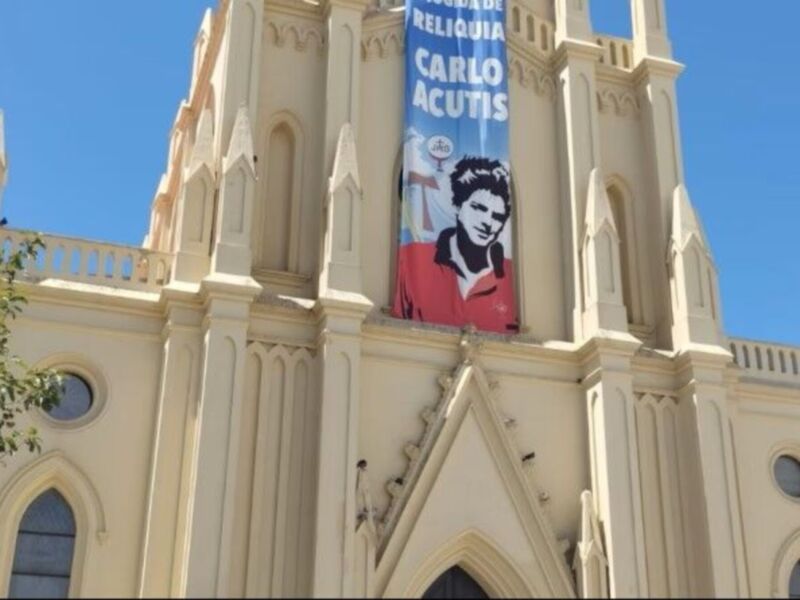 Fachada de una iglesia con detalles góticos y un cartel anunciando 'Campaña de Reliquia de Carlo Acutis'.