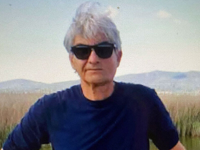 Una persona con cabello blanco y gafas de sol, posando frente a un paisaje con montañas y un cielo azul claro.
