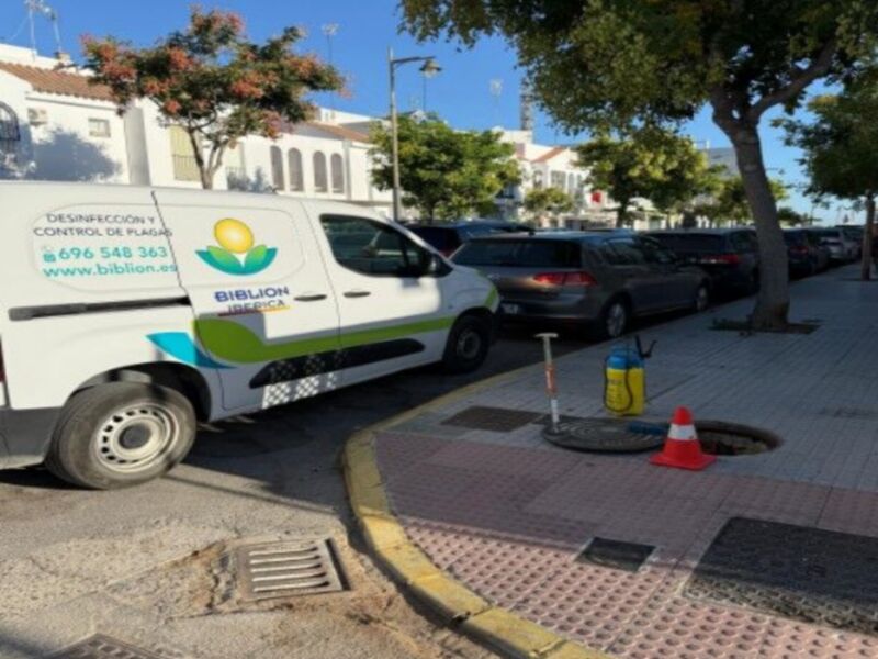 Un vehículo de desinfección y control de plagas estacionado en la acera de una calle, con un cono de seguridad en el agujero de la calle y un árbol en el fondo.