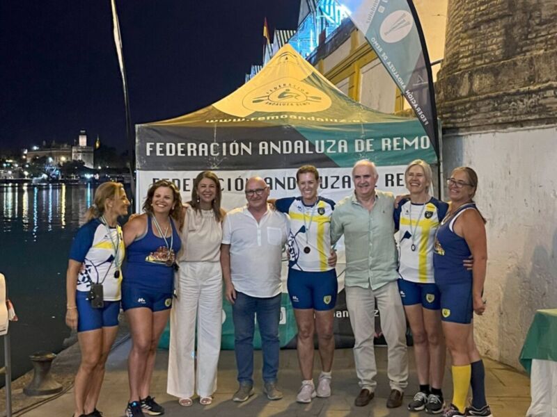 Equipo de remo celebrando una victoria en el Campeonato Andaluz de Remo, posando frente a un stand de la Federación Andaluza de Remo, con el río y la torre de la Giralda al fondo en la noche de Sevilla.