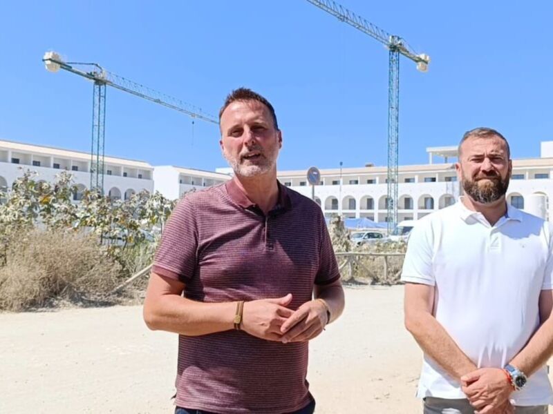 Dos hombres posan frente a una construcción en construcción, con un edificio blanco y un gran edificio en el fondo. El cielo es azul claro y hay un poste de luz en el lado izquierdo de la imagen.