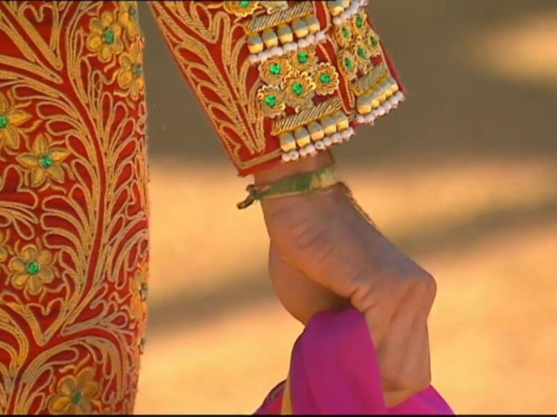 Detalle de una vestimenta tradicional con bordado dorado y flores verdes, llevada por una persona en un entorno desértico.