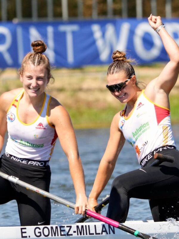 Dos mujeres compiten en una competición de remo en un río, con una bandera azul en el fondo. Ambas llevan camisetas con logos y pantalones negros de Nike Pro. La mujer en el centro levanta el brazo en victoria mientras la otra la apoya en el remo.