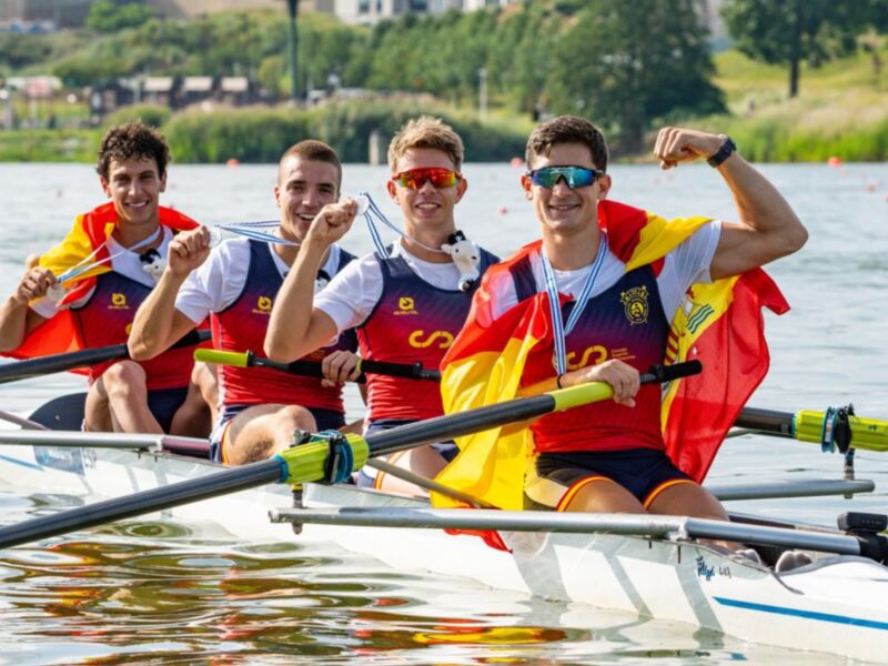 Tres deportistas celebrándose en una embarcación de remo, con banderas nacionales españolas y medallas de oro en la mano. El agua es cristalina y el paisaje es verde y tranquilo.