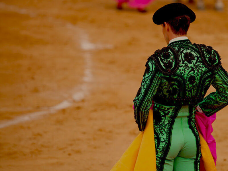Un torero con traje verde y sombrero negro en un campo de arena, mirando hacia el frente.