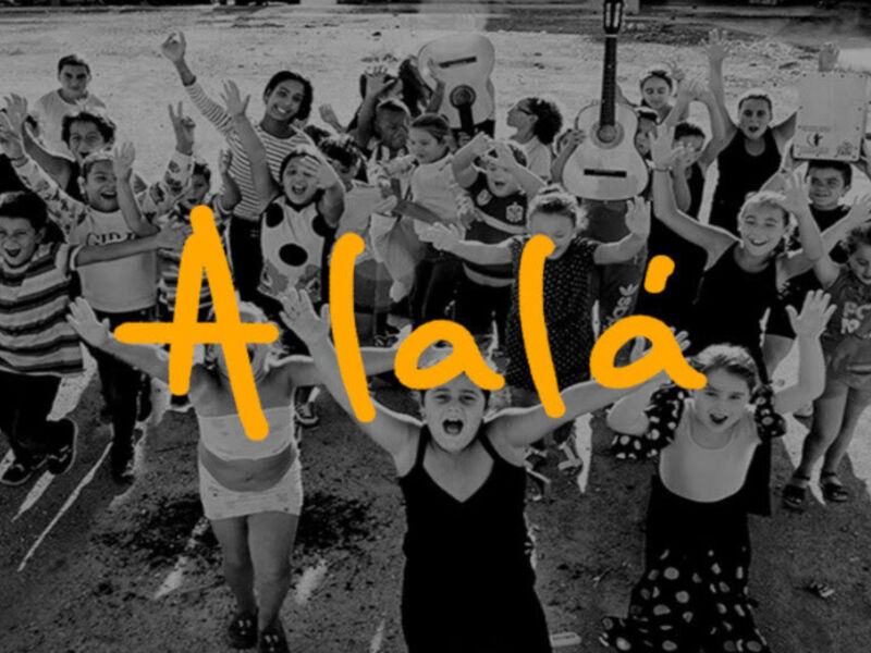 Una diversa agrupación de personas en una playa, con varios personajes y instrumentos musicales, celebrando con las manos levantadas. El texto 'Alalá' se encuentra en el centro de la imagen, destacando su importancia.