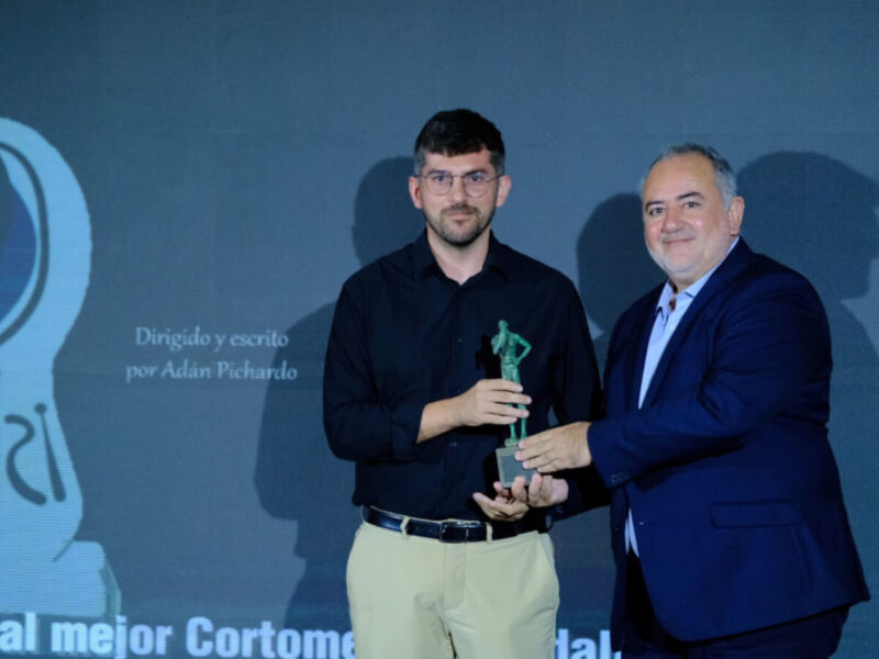 Un hombre en traje sostiene un premio mientras otro hombre lo reconoce, ambos sonríen. En el fondo, se ve un cartel con texto sobre un cortometraje ganador en la categoría de Mejor Cortometraje Nacional.