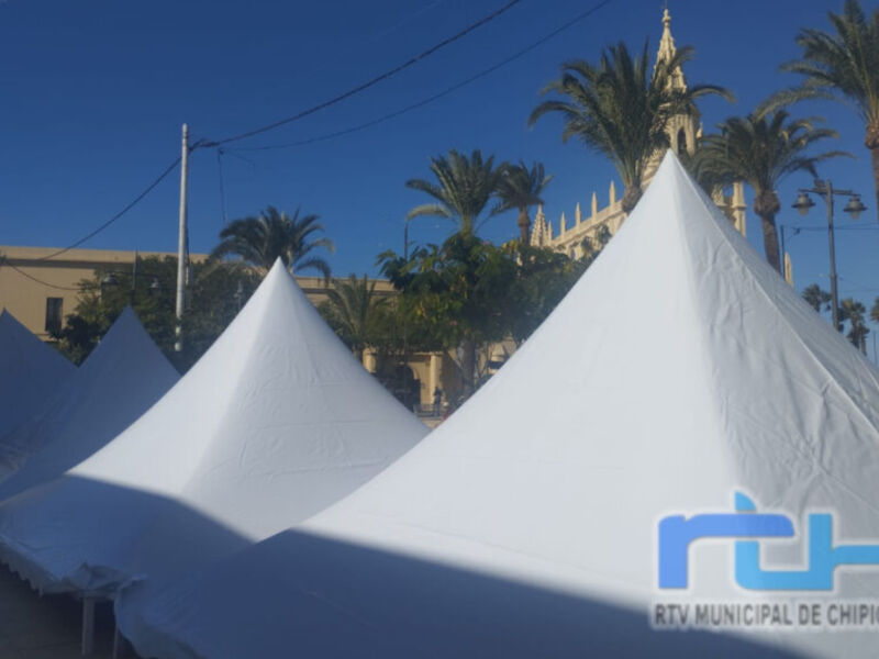 Toldos blancos en una plaza con palmeras y un edificio histórico al fondo, en Chipiona. RTV Municipal de Chipiona.