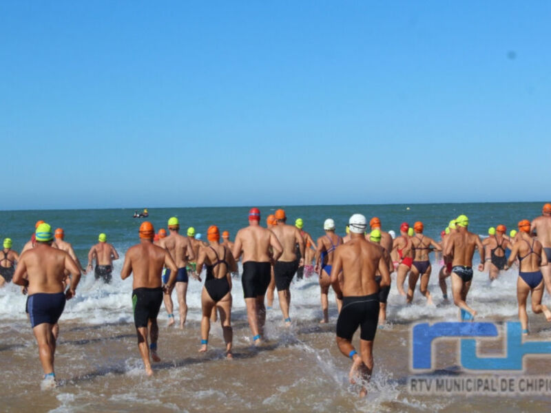Una carrera de natación en la playa, con nadadores corriendo hacia el agua. El cielo es azul y claro.