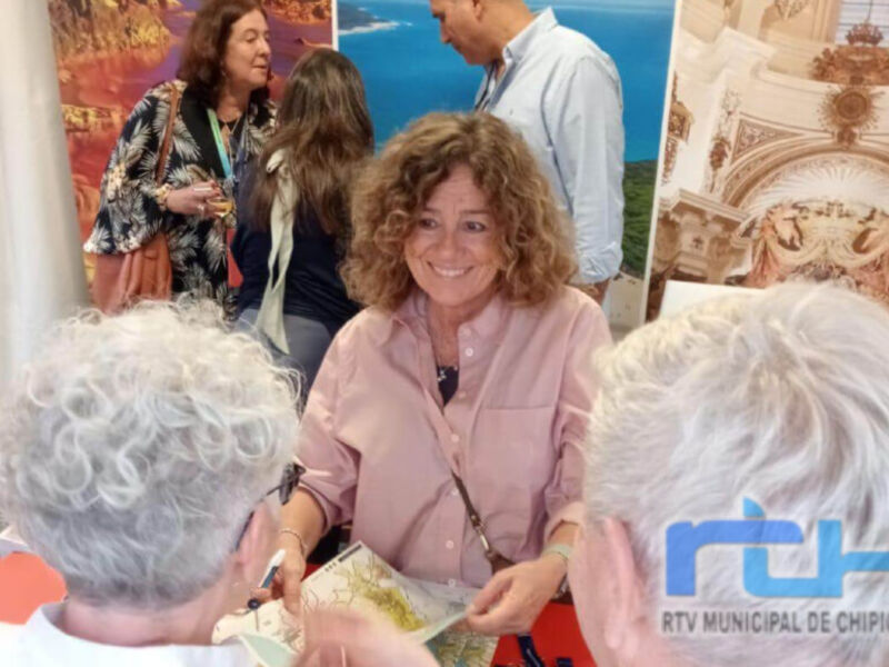 Una mujer sonriende mientras habla con otra en un evento público, con otros asistentes al fondo. En la parte inferior derecha se muestra el logo de RTV Municipal de Chipiona.