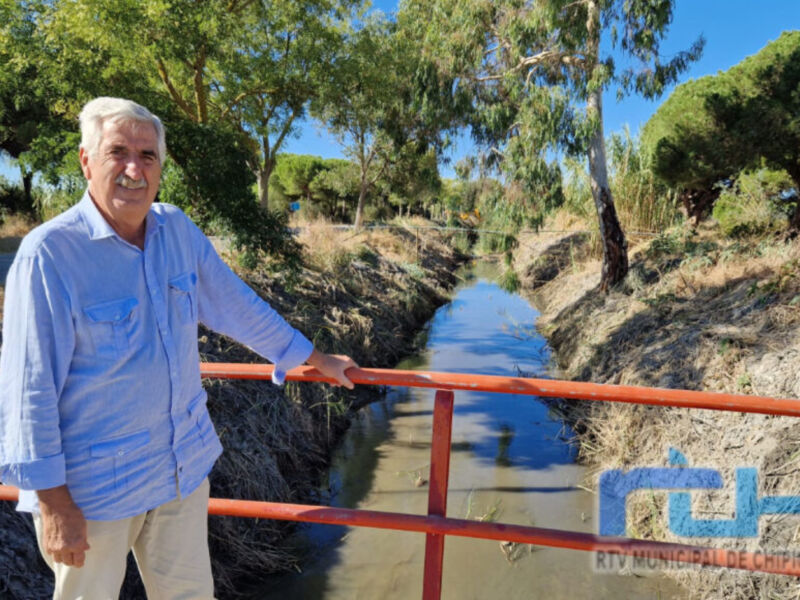 Un hombre en una camisa azul se encuentra en un puente rojo junto a un canal, con árboles y cielo azul al fondo.