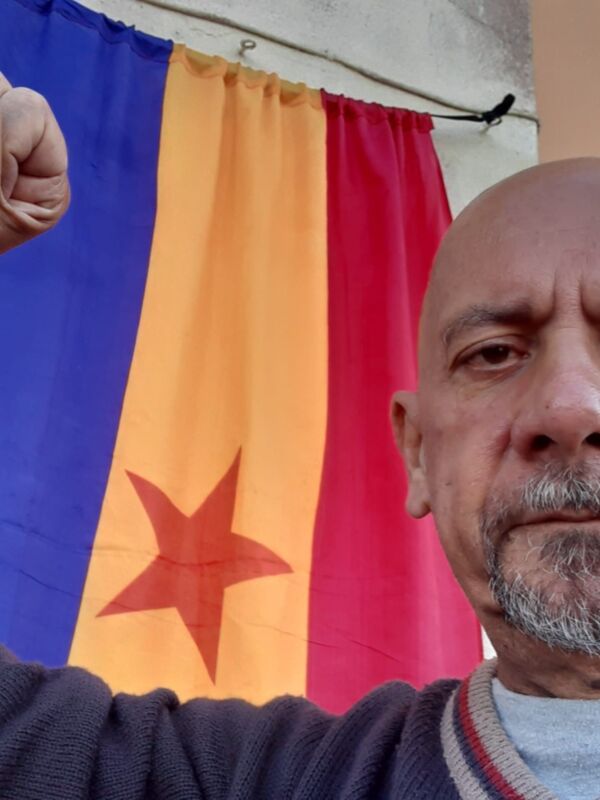 Un hombre con barba gris levanta un puño en frente de una bandera con tres franjas azul, naranja y roja, y un estrella roja en el centro.
